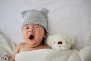 Gähnendes Baby im Bett mit Mütze und Teddy